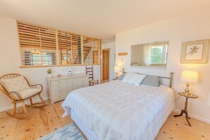 Ein Schlafzimmer mit einem großen Bett, einem Schaukelstuhl, einem Schminktisch, einem Spiegel und einem Fenster mit Jalousien.