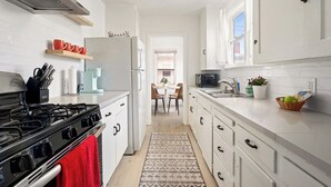 Una cucina moderna con mobili bianchi, piano cottura a gas, forno a microonde e macchina da caffè. Si vede un'area pranzo con tavolo e sedie attraverso una porta aperta.