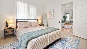Una camera da letto con un letto, comodini, uno specchio, una finestra con tende, una porta e vista su un'altra stanza.