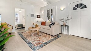 Un soggiorno moderno con un divano color beige, un tavolino rotondo e un tappeto fantasia. Si nota una porta con una finestra decorativa sulla destra e una lampada fissata al muro.