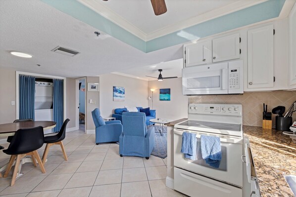 A modern kitchen with white cabinets, a microwave, and an oven. There is a dining area with a table and chairs, and a living area with a sofa and a coffee table.