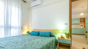 A bedroom with a bed, bedside tables, a nightstand, a window with curtains, and a view of a kitchen area.
