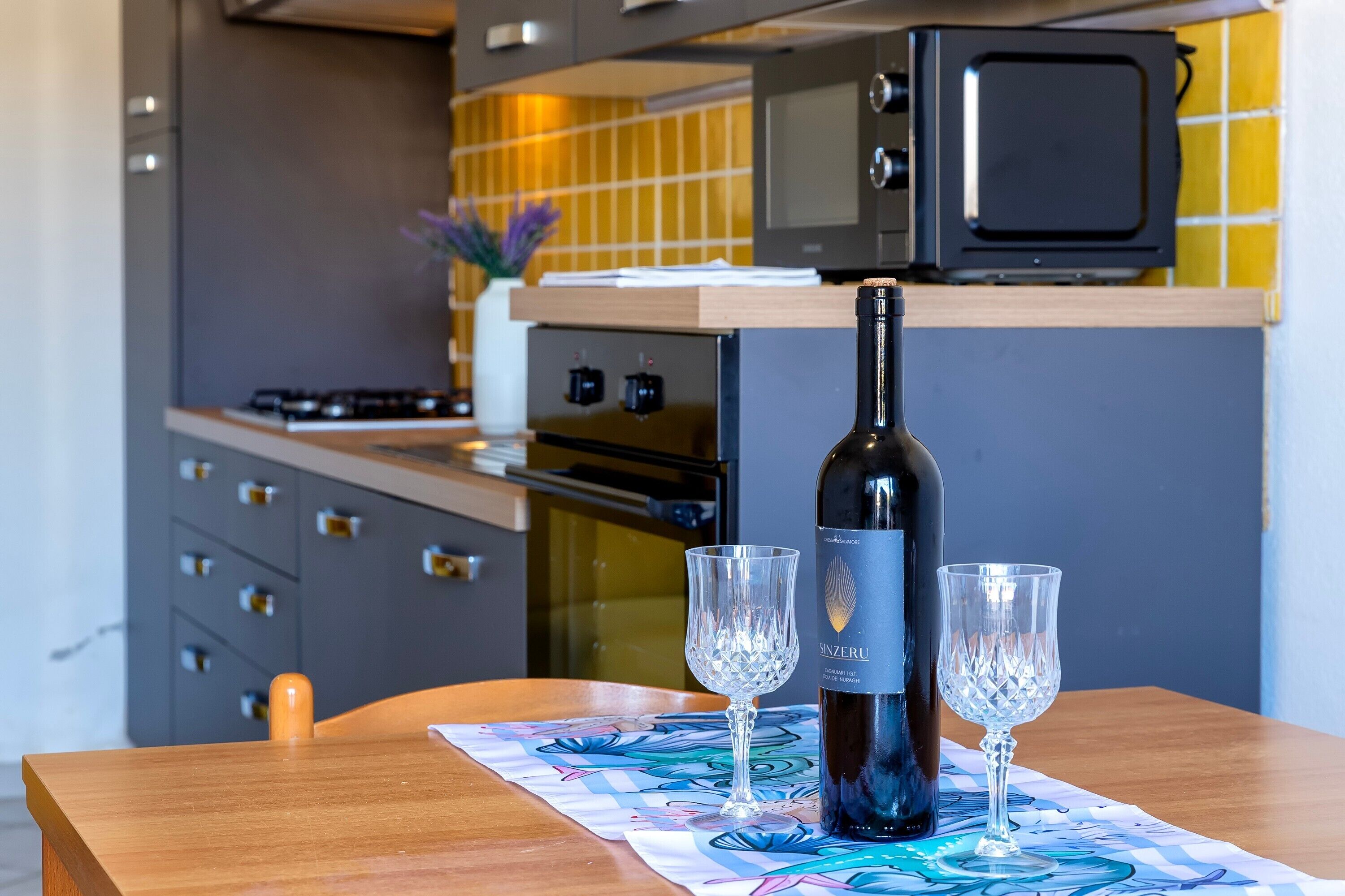 Een fles wijn en twee glaasjes op een tafel in een moderne keuken met een magnetron en oven.