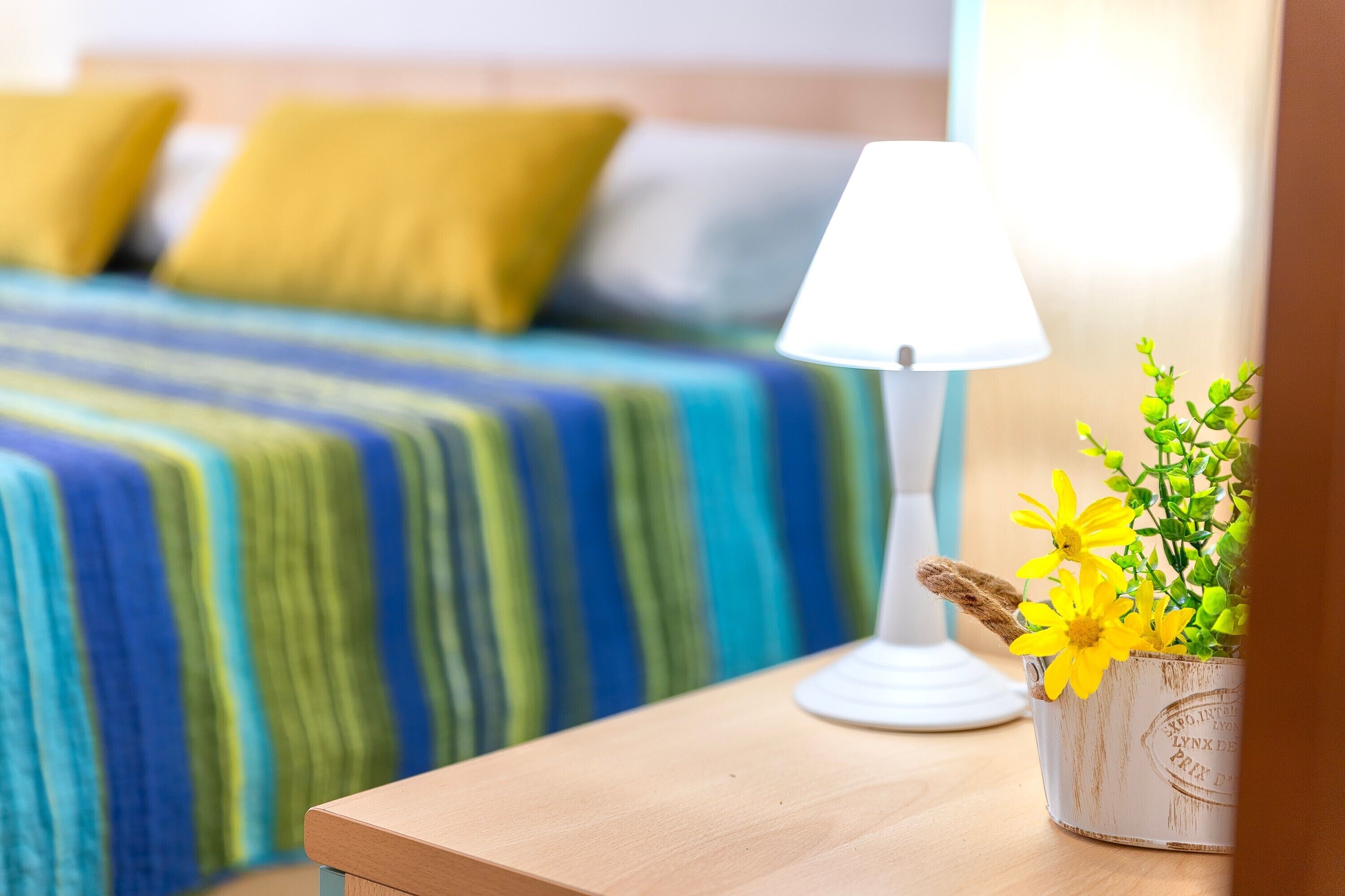 A bed with a striped blanket, a bedside table with a lamp, and a potted plant with yellow flowers.