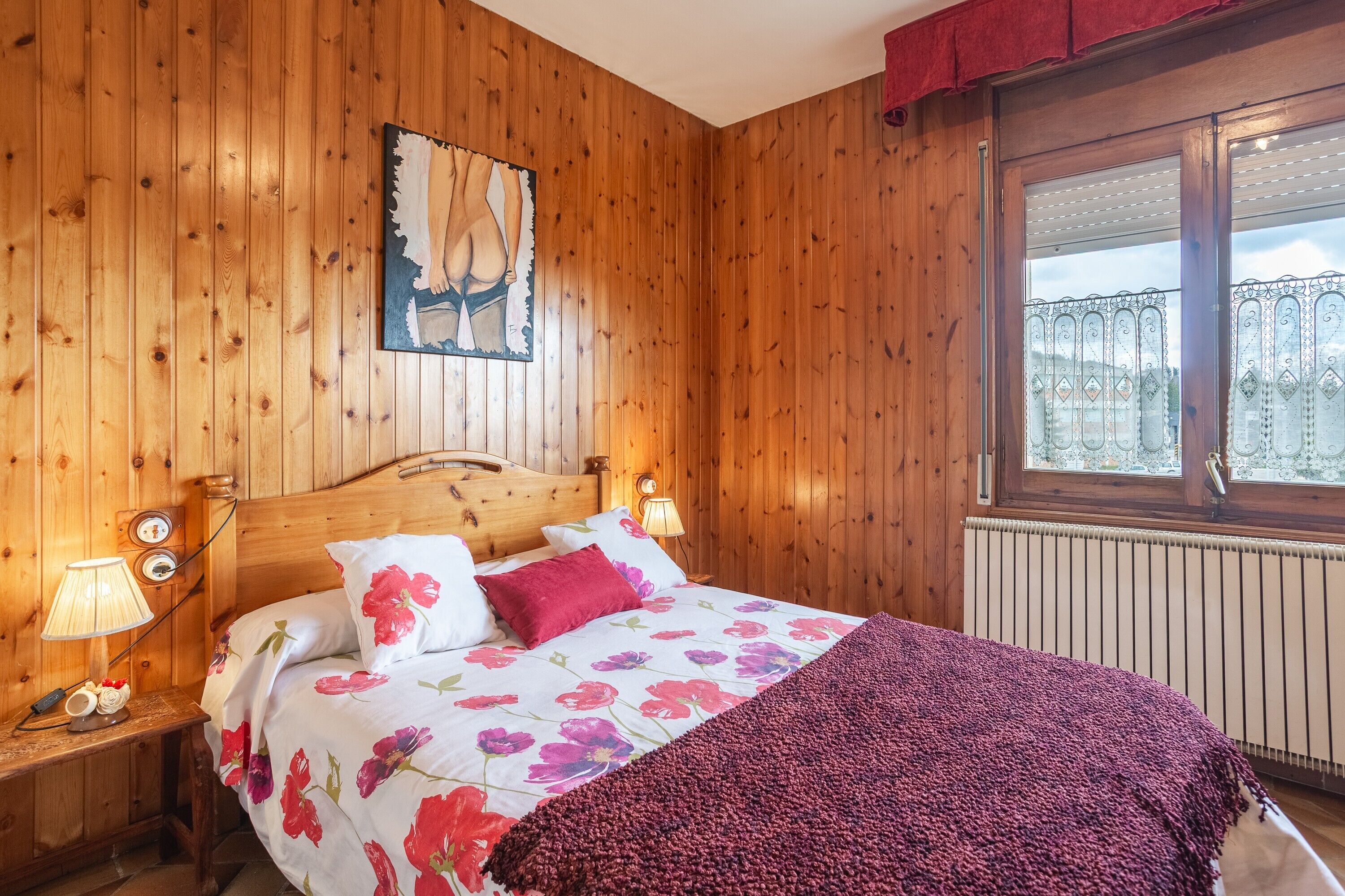 A wooden-paneled room with a bed, a nightstand, a lamp, a window with a view, and a painting on the wall.