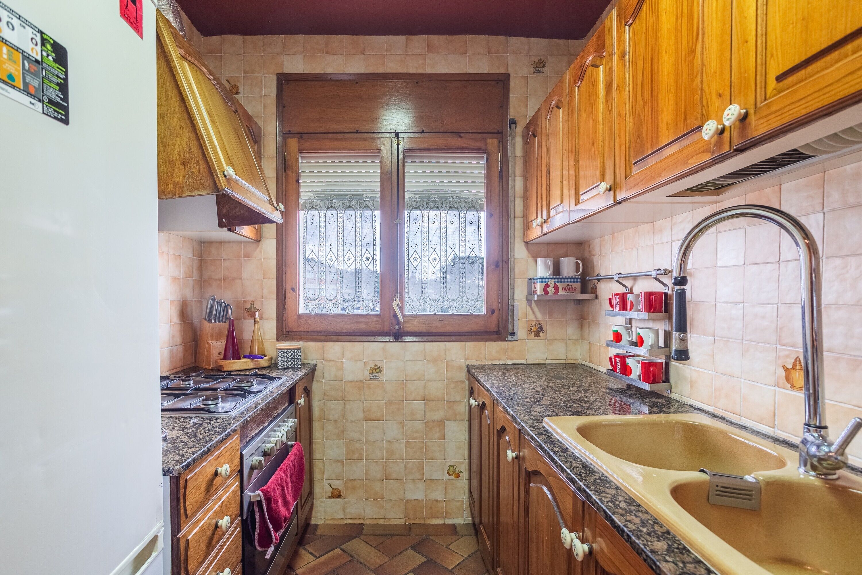 Una cocina compacta con armarios de madera, un lavamanos de acero inoxidable y una ventana con cortinas decorativas.