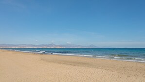 Una platja amb cel clar, mar tranquil·la i muntanyes a la distància.