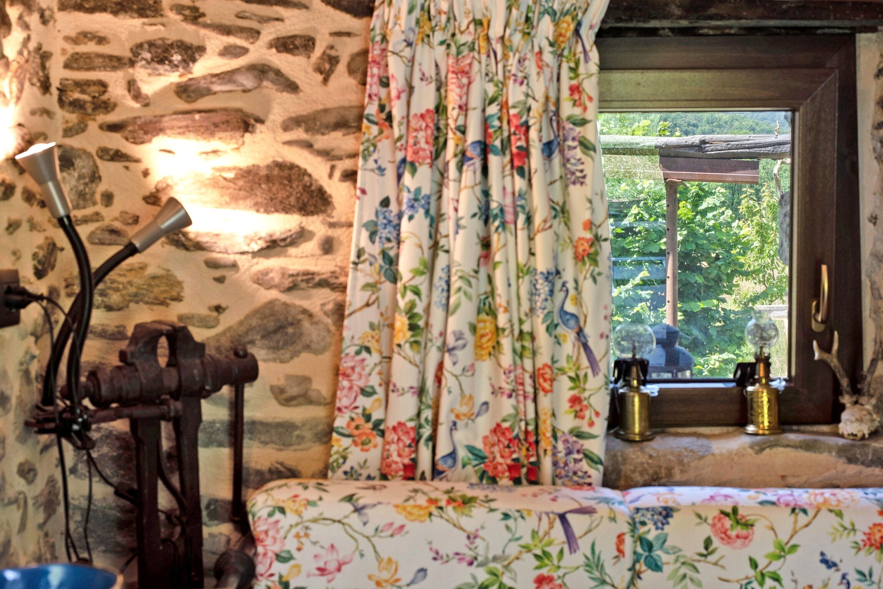 Een rustieke kamer met stenen muren, bloemen gordijnen, een vintage lamp en een raam met uitzicht op groen.