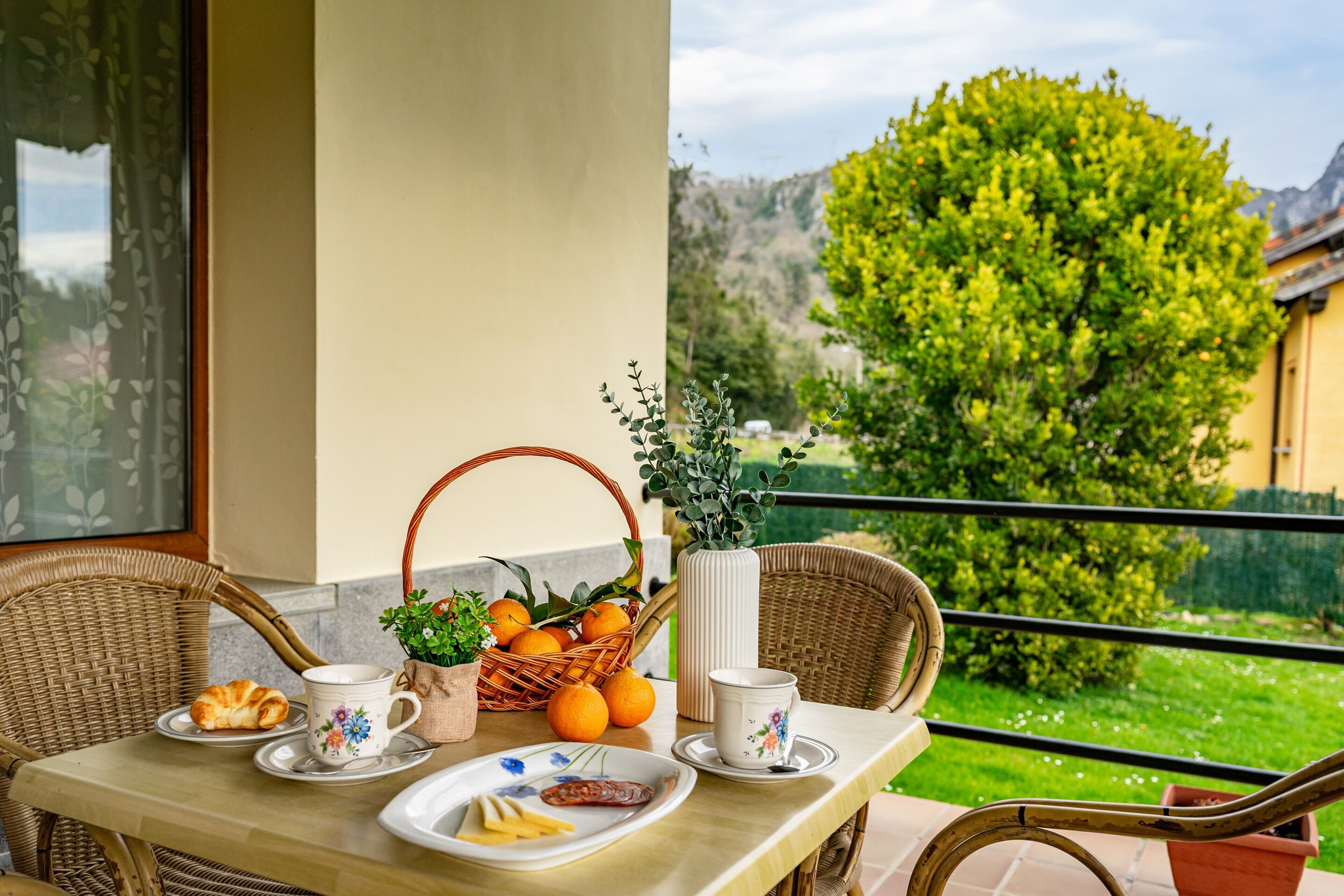 Une table à manger avec des assiettes, un panier d’oranges et un vase rempli de verdure, le tout sur un balcon offrant une vue sur un paysage verdoyant et un bâtiment.