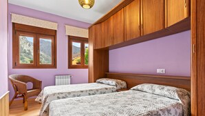 A room with two beds, wooden cabinets, a wicker chair, and large windows with a mountain view.