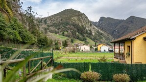 A rural landscape with a mountain, a yellow building with a porch, and a green hedge.