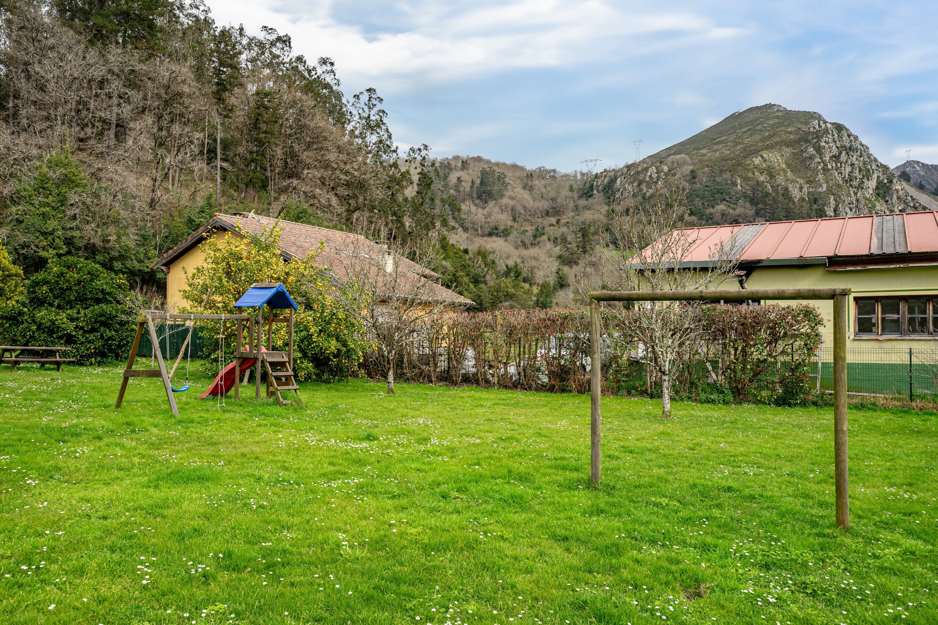 A grassy outdoor area with a swing set, a small building with a red roof, and a mountain in the background.