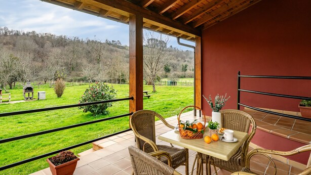 A covered patio with wicker furniture, a table with fruit and a vase, and a view of a green landscape.