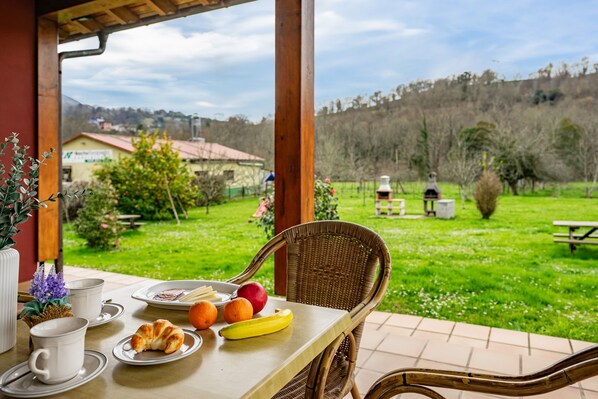 A table with breakfast items, a wicker chair, and a view of a green landscape with trees and a building in the distance.