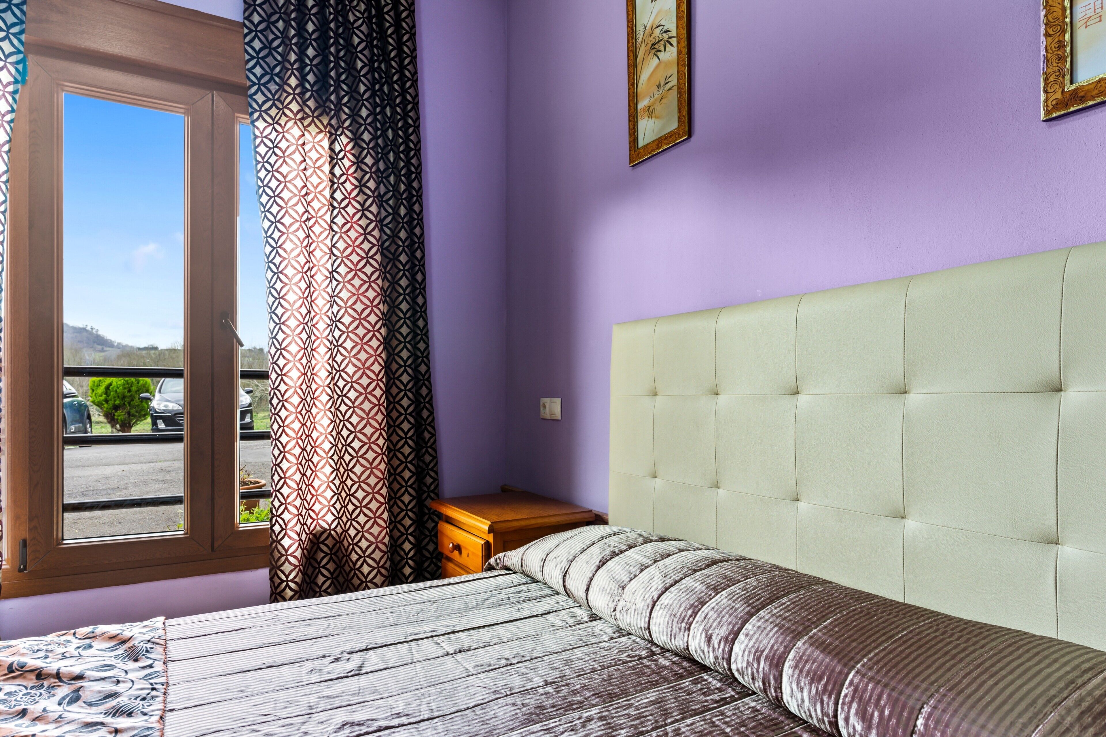 A bedroom with a bed, a nightstand, a window with curtains, and a view of the outside.