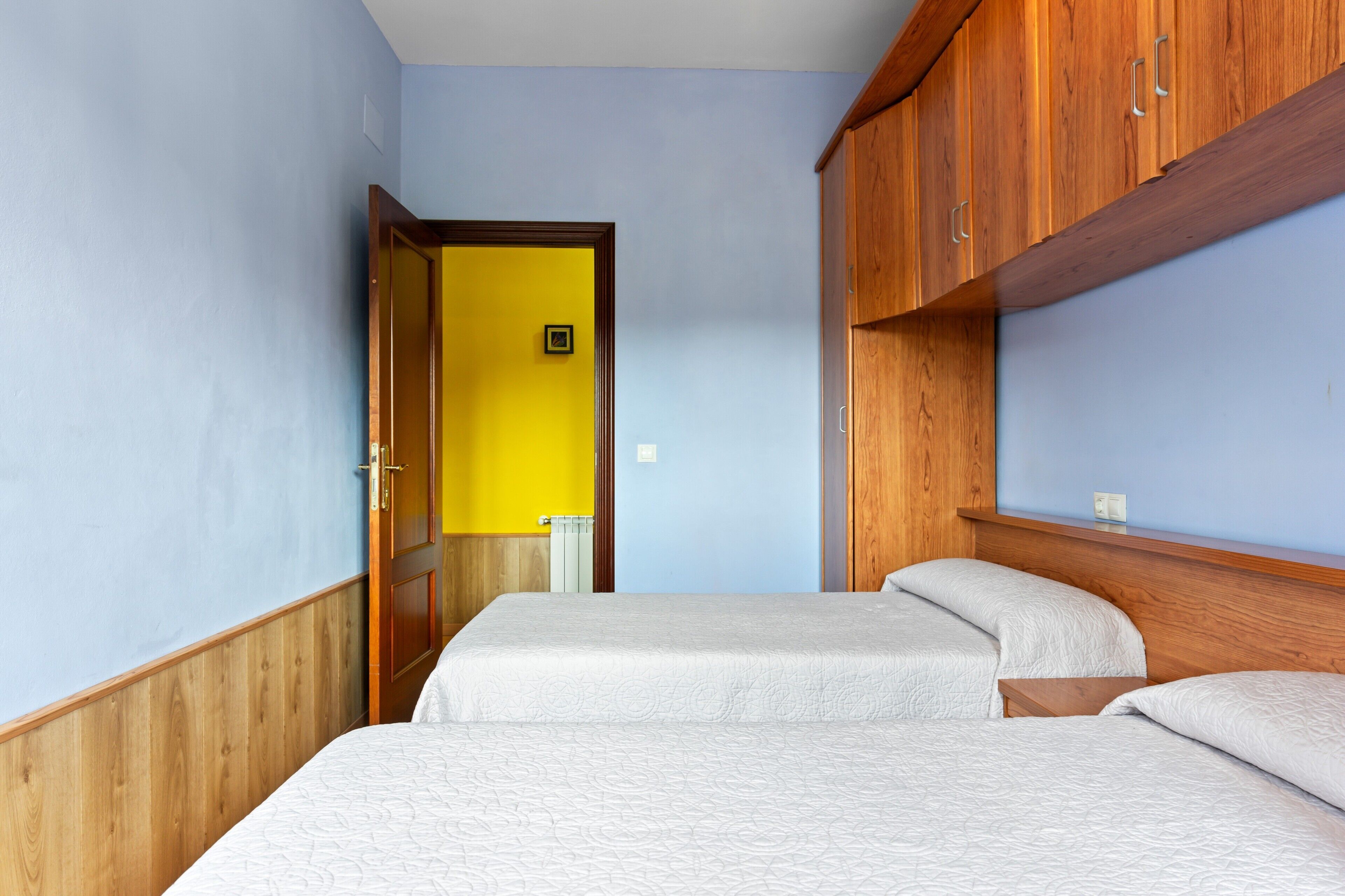 Een kamer met twee bedden, houten kasten, een gele deur en een lichtblauwe muur.