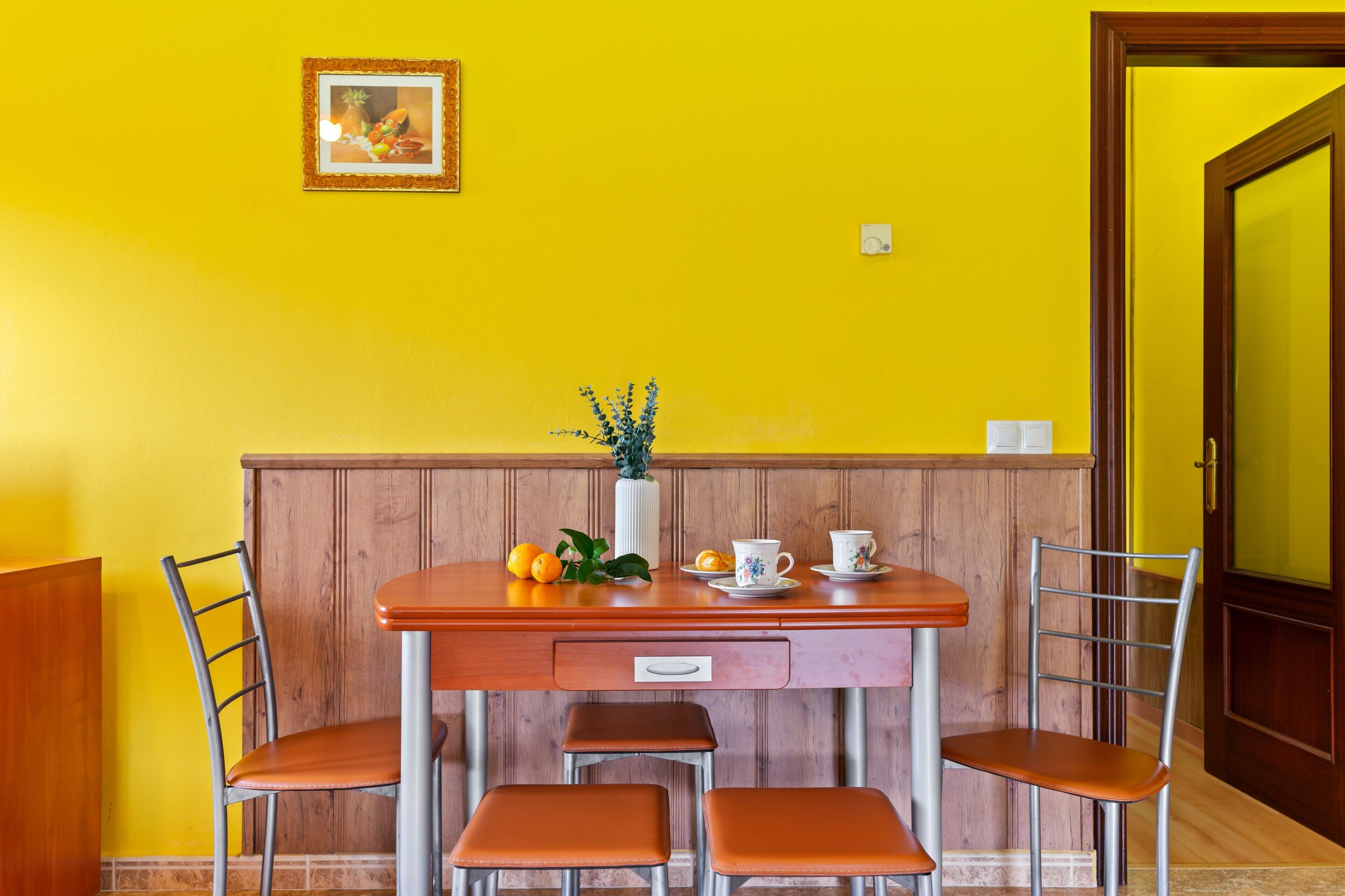 A dining area with a wooden table, four chairs, a vase with flowers, and a framed picture on the wall.