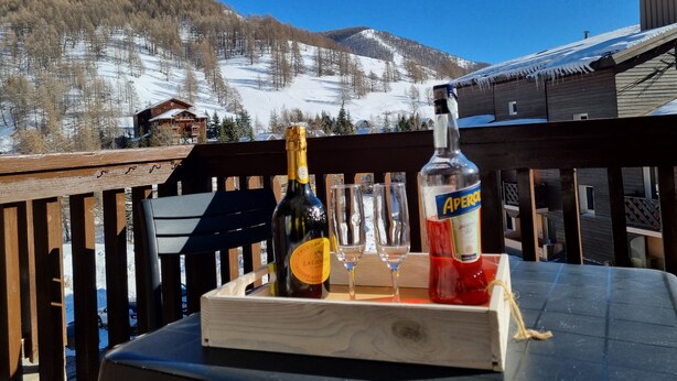 Un terrazza in legno con un tavolo, una bottiglia di Aperol e una bottiglia di vino, con vista su un paesaggio montano innevato.