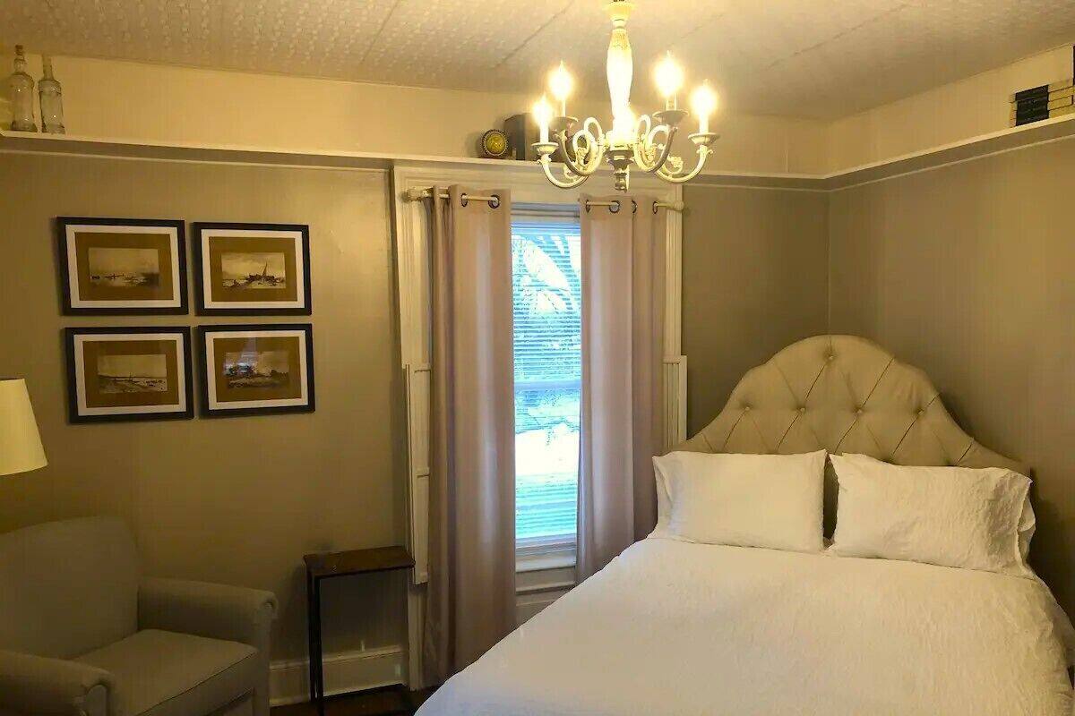 Ein Schlafzimmer mit einem großen Bett, einem Sessel, einem Nachttisch, einer Chandelüster und einem Fenster mit Vorhängen.