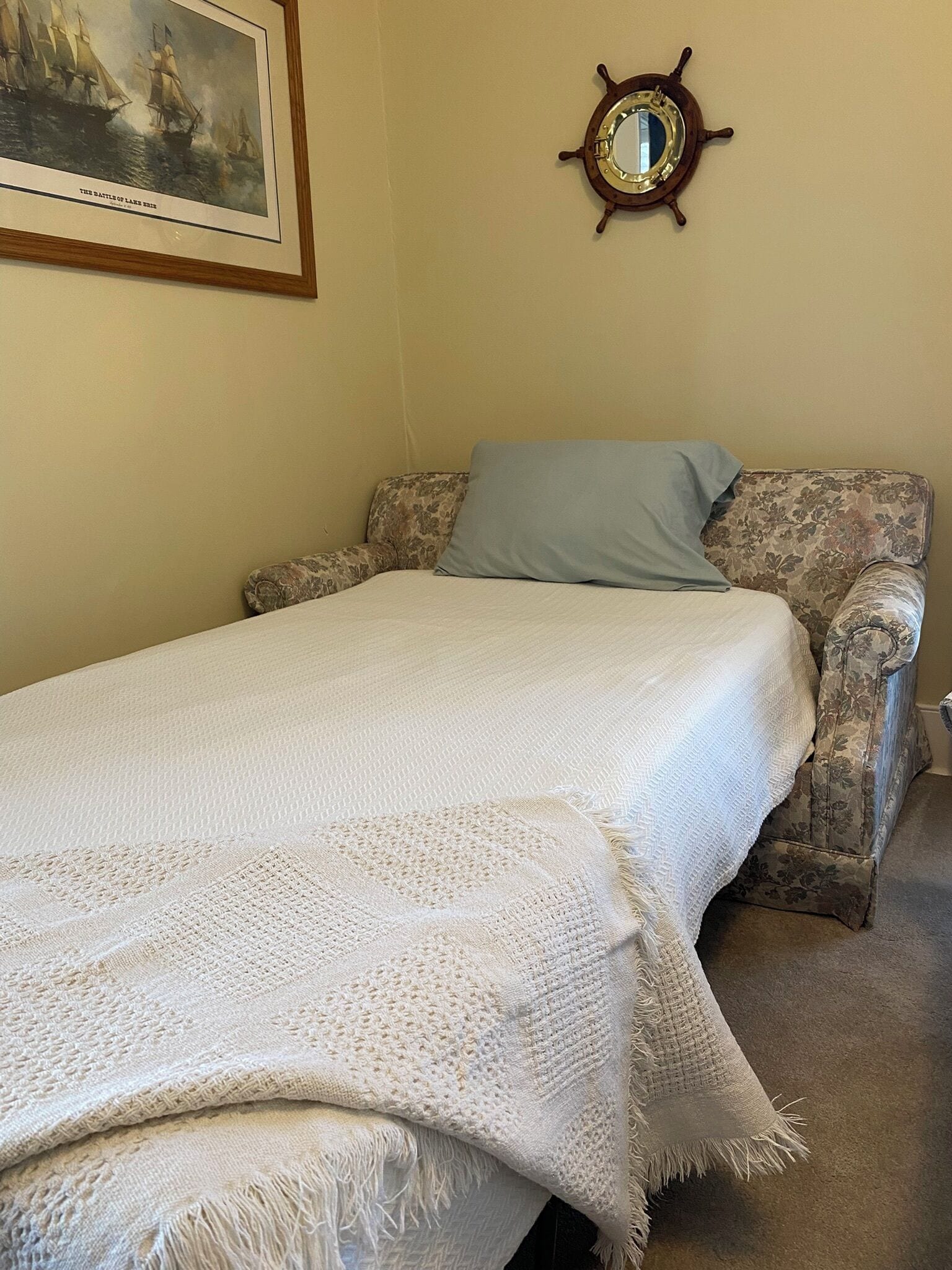 A bedroom with a bed, a floral-patterned armchair, a framed painting, and a ship's wheel decoration.