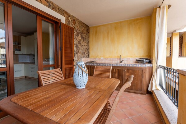 A wooden dining table with chairs, a vase, a kitchen with a stone wall, and a balcony with a railing.