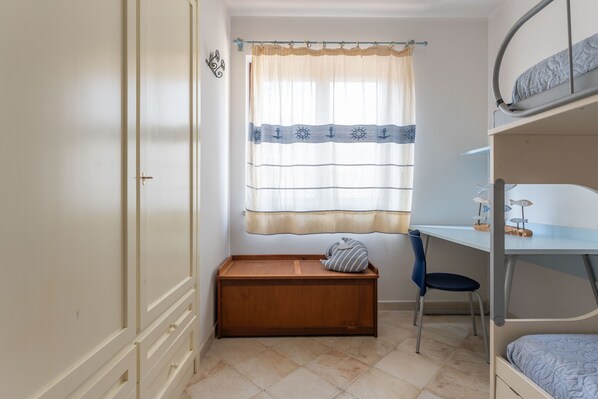 Une chambre avec un lit superposé, un bureau avec une chaise, un petit banc en bois et une fenêtre avec un rideau.
