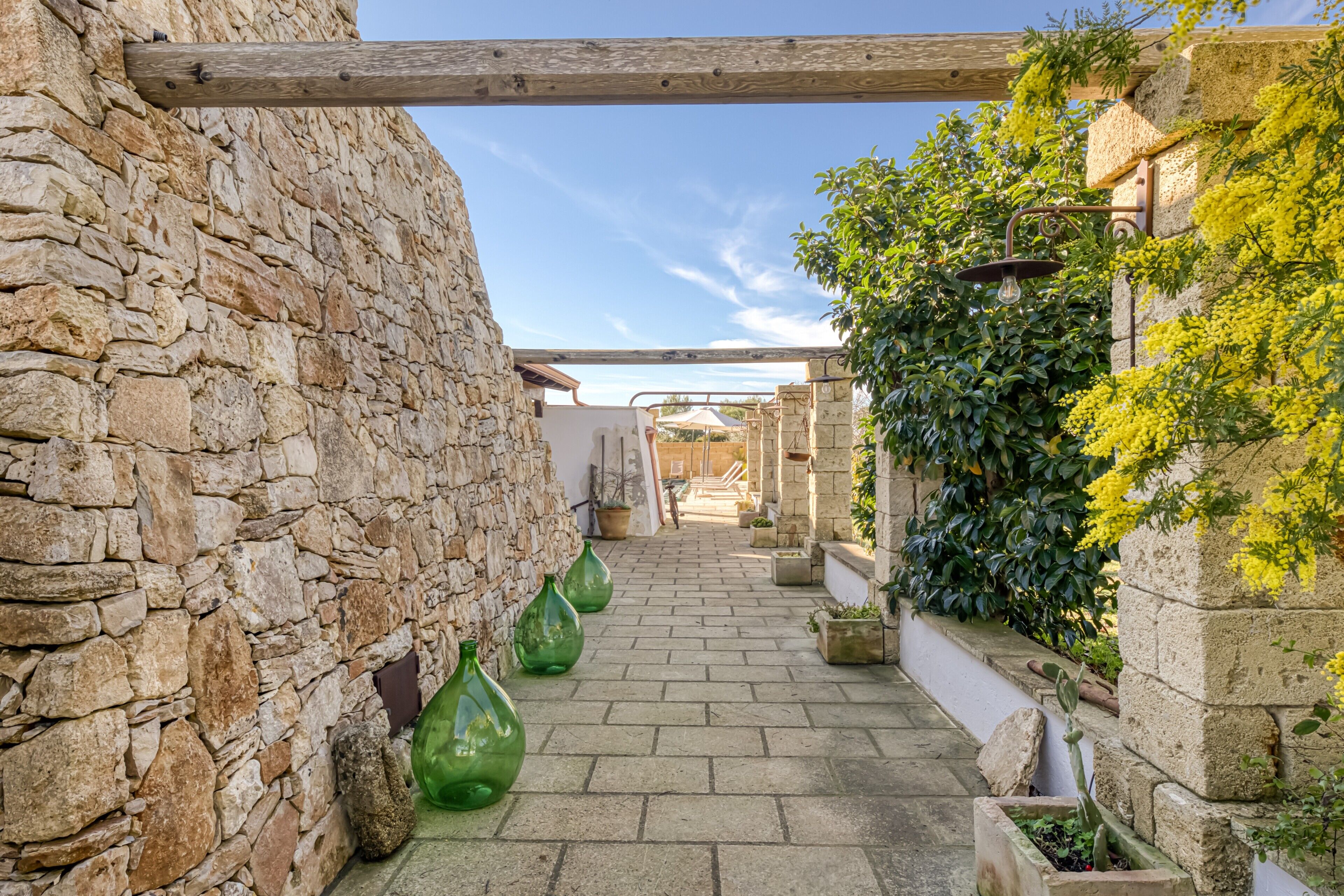Une ruelle aux murs de pierre, surmontée d’une poutre en bois, agrémentée de plantes en pot et offrant une vue sur un ciel dégagé.
