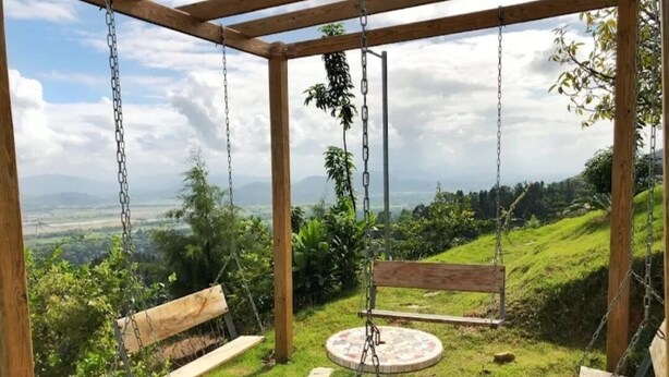 Une balançoire en bois, avec des bancs et une table, installée devant une colline verdoyante et des montagnes lointaines.