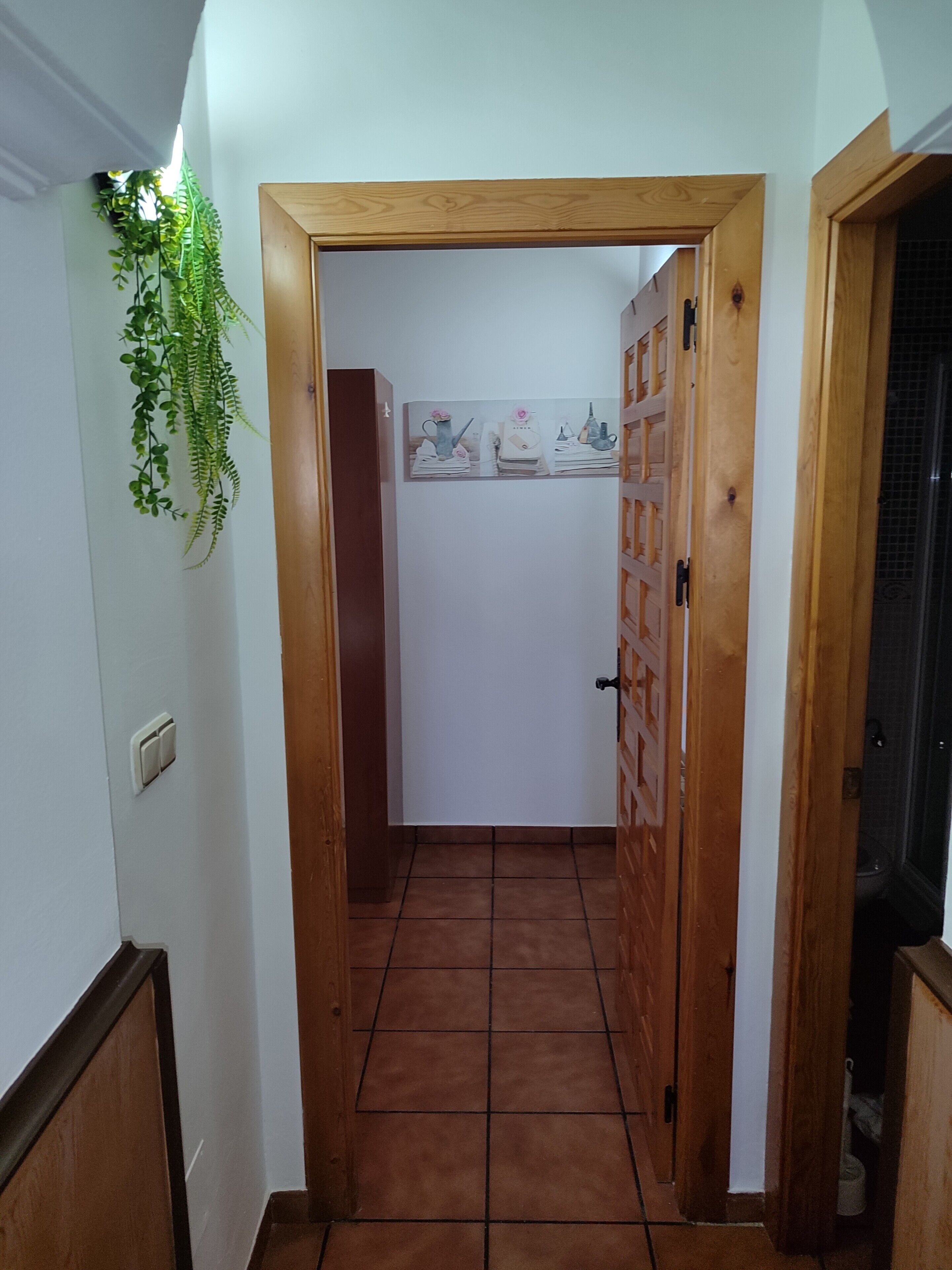 Un couloir étroit doté d’un cadre de porte en bois, d’un sol carrelé et d’une étagère fixée au mur, sur laquelle sont disposés des objets décoratifs.