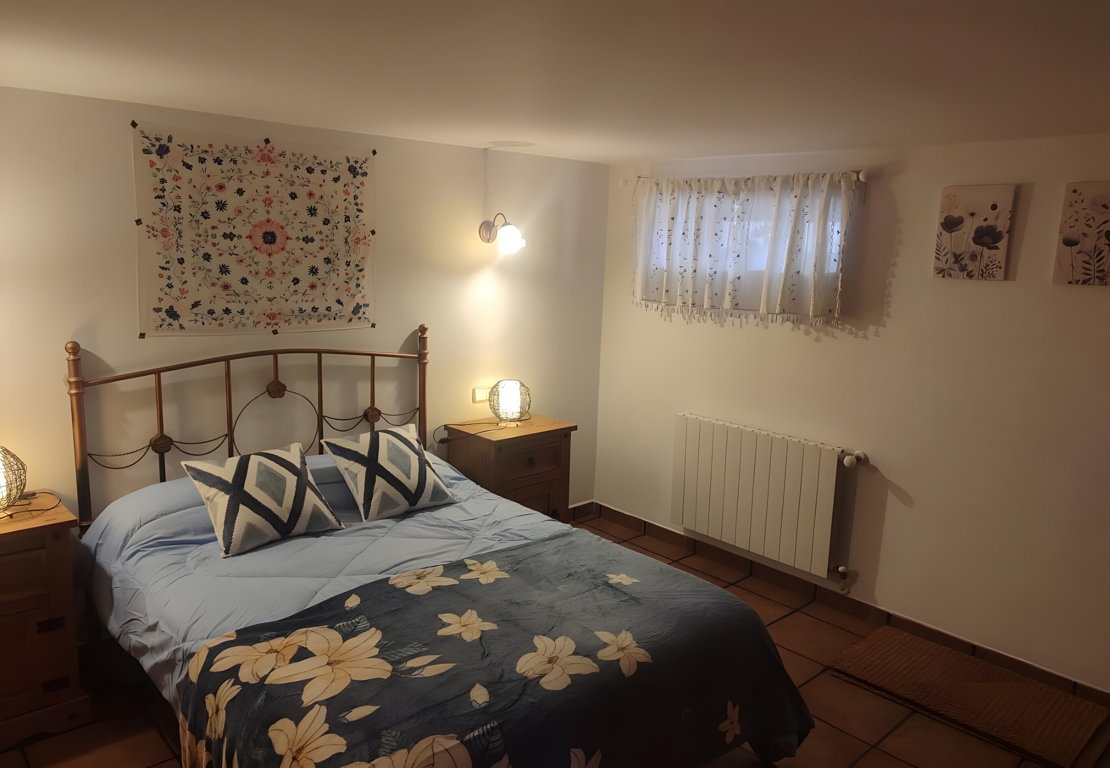 A bedroom with a bed, nightstands, a wall-mounted lamp, a radiator, and a window with curtains.