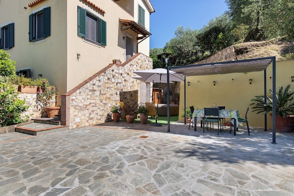 Un patio con suelo de piedra, mobiliario exterior, una zona de estar cubierta y un edificio con contraventanas verdes.