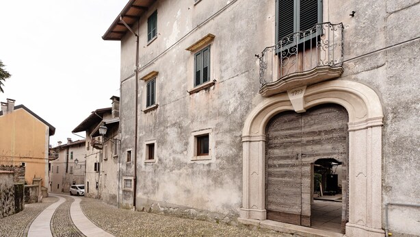 A narrow cobblestone street with a building featuring a large arched doorway and a balcony with wrought iron railings.