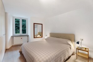 Une chambre à coucher avec un lit, une table de chevet avec une lampe, une fenêtre donnant sur des arbres et un radiateur.