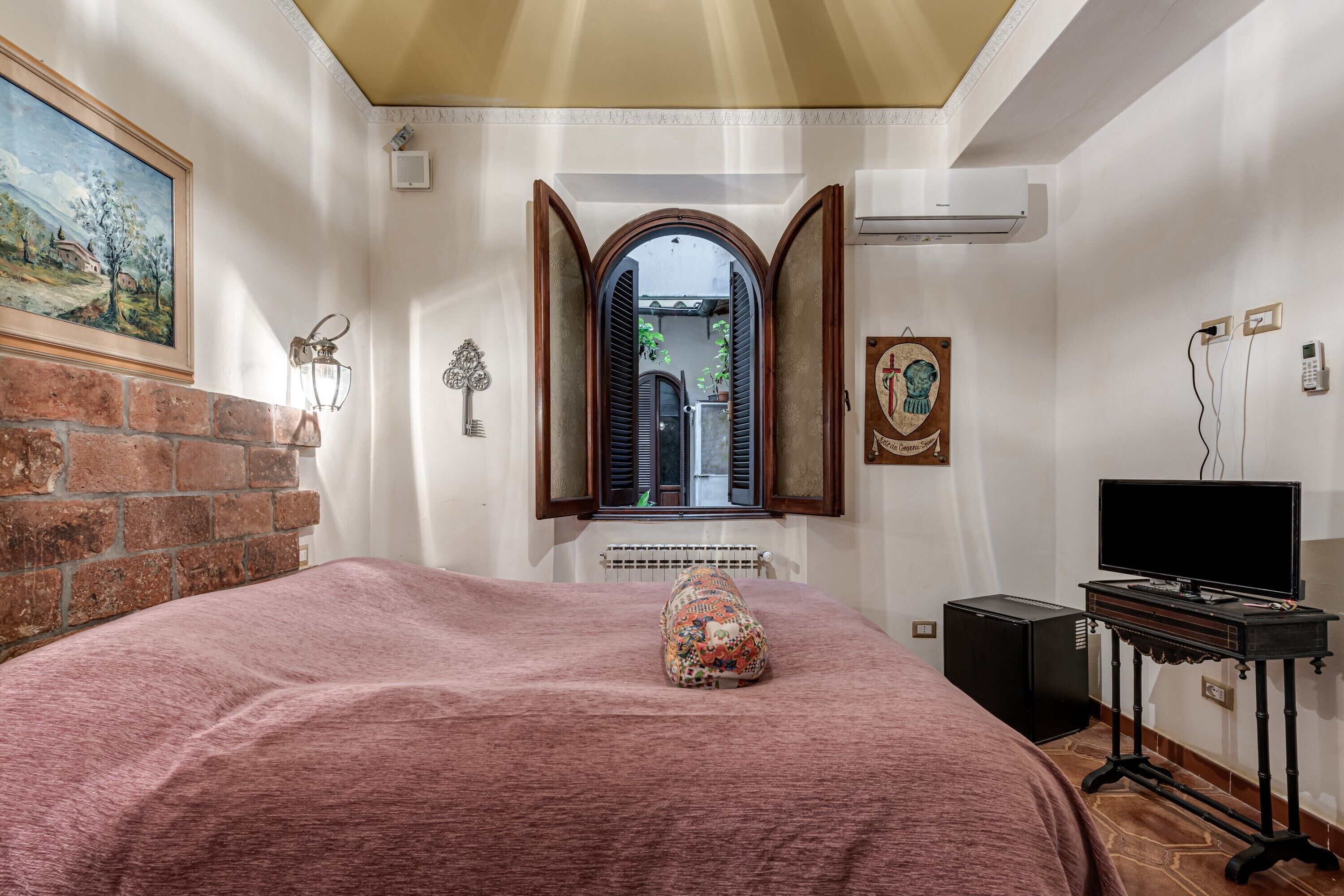 A bedroom with a bed, a television on a stand, a brick wall, a window with shutters, and a painting on the wall.