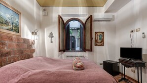 A bedroom with a bed, a television on a stand, a brick wall, a window with shutters, and a painting on the wall. - Room 'Matroniale 3' with Wi-Fi and Air Conditioning (Firenze)