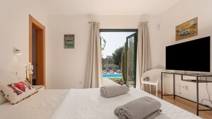 A hotel room with a bed, a desk with a TV, a view of a swimming pool, and a door leading to another room. - El Refugio Vacation Home with Mountain Views, Private Pool, and Wi-Fi (La Oliva)