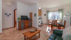 Una sala de estar con un sofá, una mesa de madera, un televisor, un área de cocina con lavadora y una mesa de comedor con sillas.