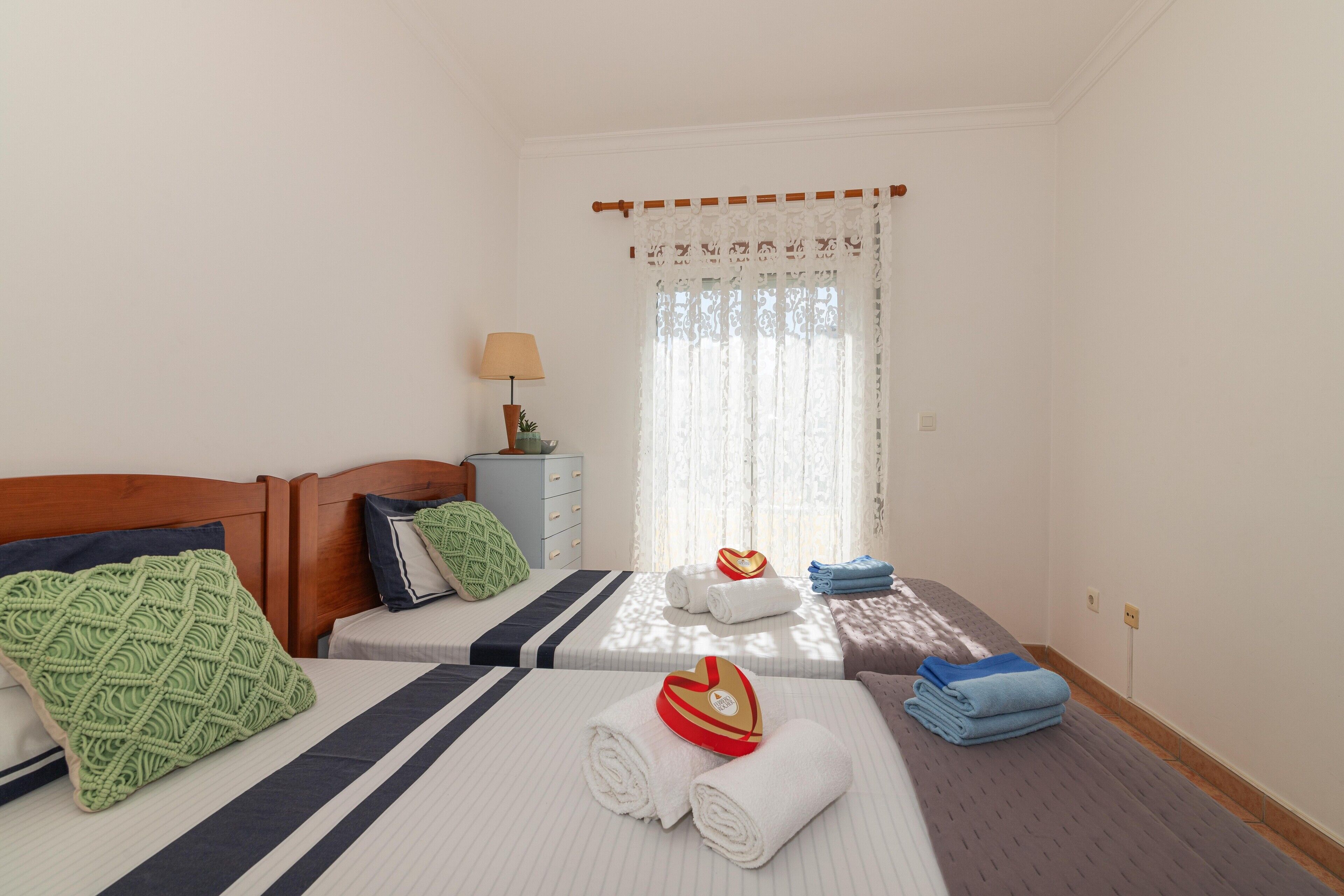 A hotel room with two beds, a nightstand, a lamp, and a window with curtains.