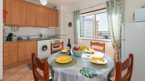 Cocina compacta con armarios de madera, lavadora, mesa de comedor con mantel verde y una ventana con cortinas.