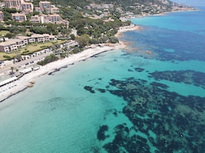 Vue aérienne d’une zone côtière avec des bâtiments, une plage et une eau d’un bleu limpide.