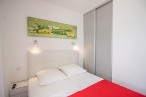 A bed with white bedding, a red accent, a nightstand with a remote, and a wall-mounted artwork. - Apartment 'Bel Neuf, Climatisé' with Sea View, Shared Pool and Wi-Fi (Albitreccia)