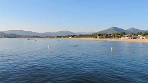 Ein ruhiger See mit Booten, einem Strand und Bergen im Hintergrund.