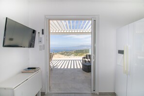 A room with a TV mounted on the wall, a door leading to a balcony with a view of the sea, and a refrigerator. - Apartment 'Sublime Neuf' with Sea View, Shared Pool and Wi-Fi (Albitreccia)