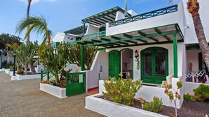 Un edificio blanco con detalles verdes, un porche cubierto y un jardín con cactus y plantas pequeñas.
