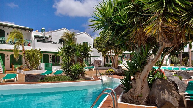 Zona de piscina con sillones de descanso, un árbol grande y un edificio con techo verde.