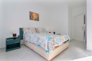 Un dormitorio con cama, mesita de noche, lámpara, un cuadro en la pared y una puerta que da a otra habitación.