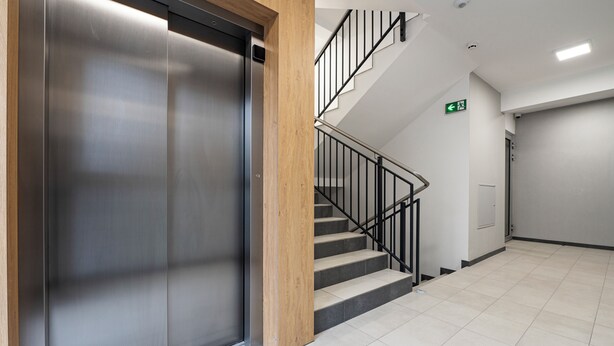 Un ascensor moderno con estructura de madera, una escalera con barandillas negras y un suelo de baldosas.