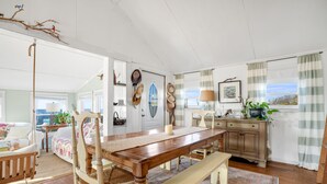 Un coin repas avec une table en bois, des chaises et un banc. Il y a une fenêtre avec des rideaux, une lampe et une plante sur la commode.