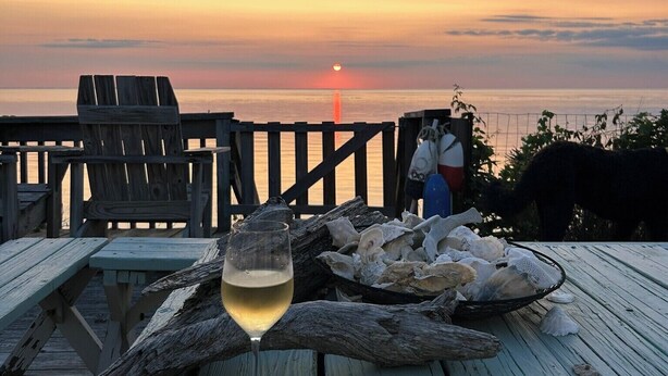 Une terrasse en bois, un verre de vin blanc, un panier d’huîtres et une vue sur le coucher de soleil.