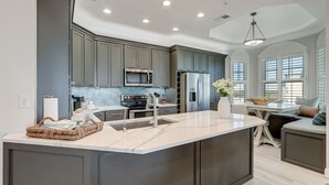 A modern kitchen with grey cabinets, a marble island, stainless steel appliances, and a dining area with a white table and chairs.